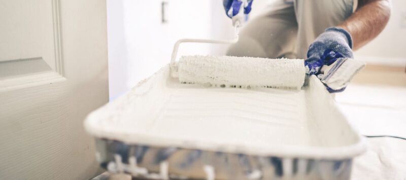 A painter dips a roller into a tray filled with white paint while wearing gloves and preparing to paint a door